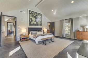A spacious and serene master bedroom at Stillson Meadow, featuring a large bed with a dark upholstered headboard, white bedding, and plush throws, under a vaulted, paneled ceiling with recessed lighting.