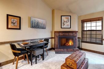 The dining area and fireplace at Copperbottom 311, featuring a rustic brick fireplace with a roaring fire, a black dining table set for two, and a window with a wooden blind.