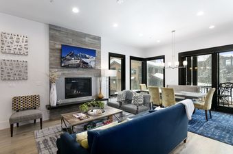 A modern and spacious living room in Juniper 901 with a large flat-screen TV above a fireplace, comfortable seating, and an integrated dining area with large windows showcasing a snowy exterior.