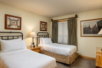 A cozy twin bedroom at Montaneros 306 features two beds with white linens, a warm lamp between them, and rustic artwork on the walls.