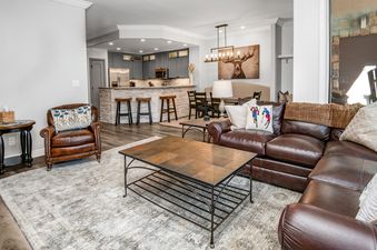 The open-concept living space features a spacious brown leather sectional sofa, a tiled coffee table, and a plush area rug, with a fully equipped kitchen and dining area visible in the background.