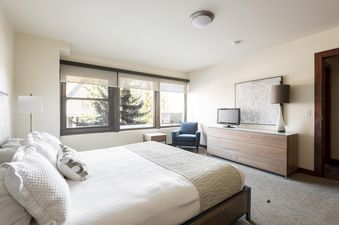 The bedroom at Lowell 201B features a large bed with white bedding, a wooden dresser with a television, and windows overlooking an outdoor scene.