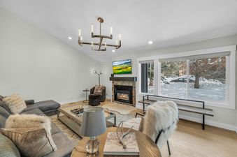 A cozy living room at Last Run Luxury features a large fireplace, comfortable seating, and a large window showcasing a snowy landscape.