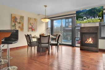 A dining area with a glass-top table, four chairs, a breakfast bar, and a fireplace, with a television displaying a mountain scene.