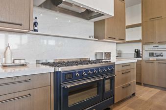 A modern kitchen with a large blue range stove, white marble countertops and backsplash, and light wood cabinetry.