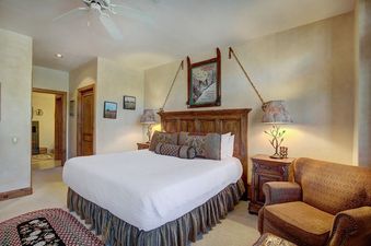 The bedroom at Arrowhead Village Condominium Premium 3 Bedroom features a king-sized bed with a rich wooden headboard, flanked by two nightstands with lamps, and decorated with a unique ski-themed artwork above the bed.