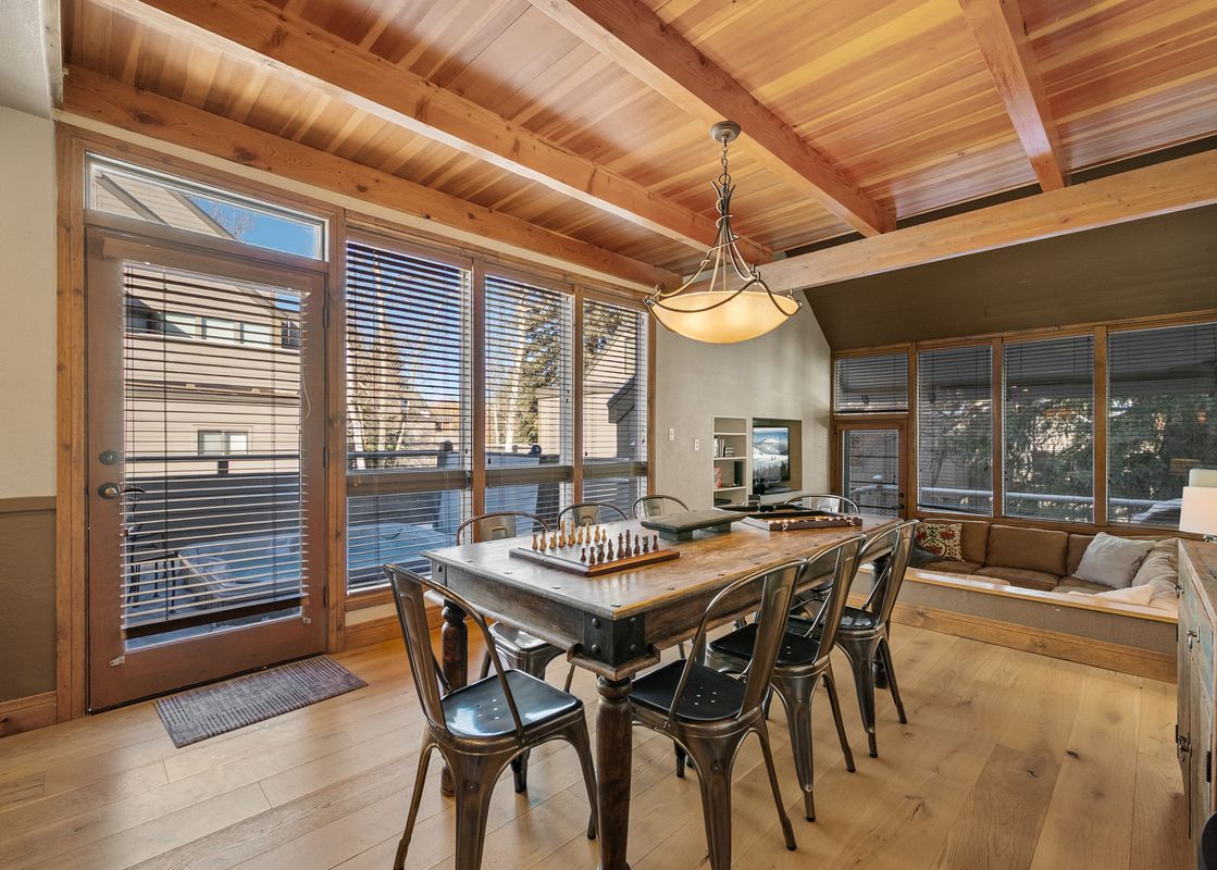 The dining area at Three Kings Powder Lodge features a large wooden table with a chess set, surrounded by metal chairs, illuminated by a chandelier and natural light from expansive windows and a glass door.