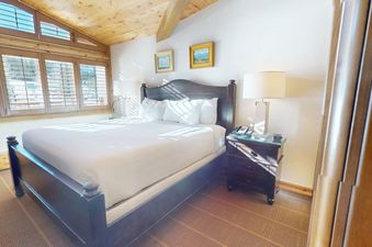 The bedroom in Manor Vail 227 features a large, comfortable bed with white linens, a wooden headboard, and a bedside table with a lamp. Sunlight streams in through the large windows.