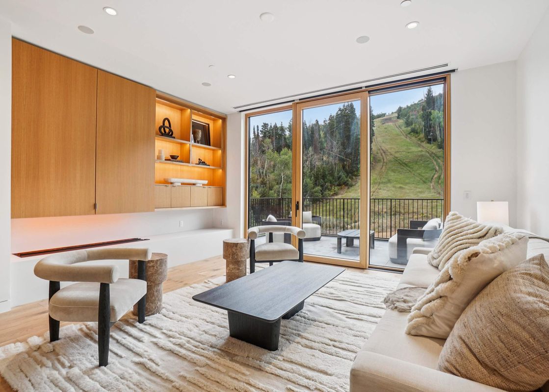 The Founders Place 2401 living area features a cream sofa, modern armchairs, and a sleek coffee table, with a built-in wooden cabinet and large glass doors leading to a balcony with mountain views.