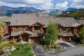 An aerial view of the TRISTANT 115, a large log cabin-style house with a wooden shake roof, nestled into a hillside with vibrant green and autumnal trees, set against a dramatic backdrop of towering, rugged mountains under a partly cloudy sky.