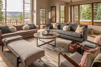 A spacious and modern living room featuring two large sofas, a wooden coffee table, and expansive windows offering views of mountains and a hot tub.