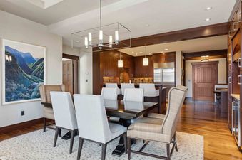 A dining room with a wooden table and chairs, a large picture on the wall, and a doorway leading into the kitchen.