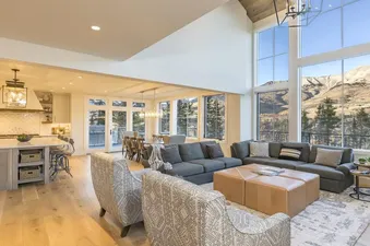 A spacious and modern open-concept living area in Overlook Haus with a gray sectional, patterned armchairs, and a large tan leather ottoman, offering expansive mountain views through floor-toceiling windows.