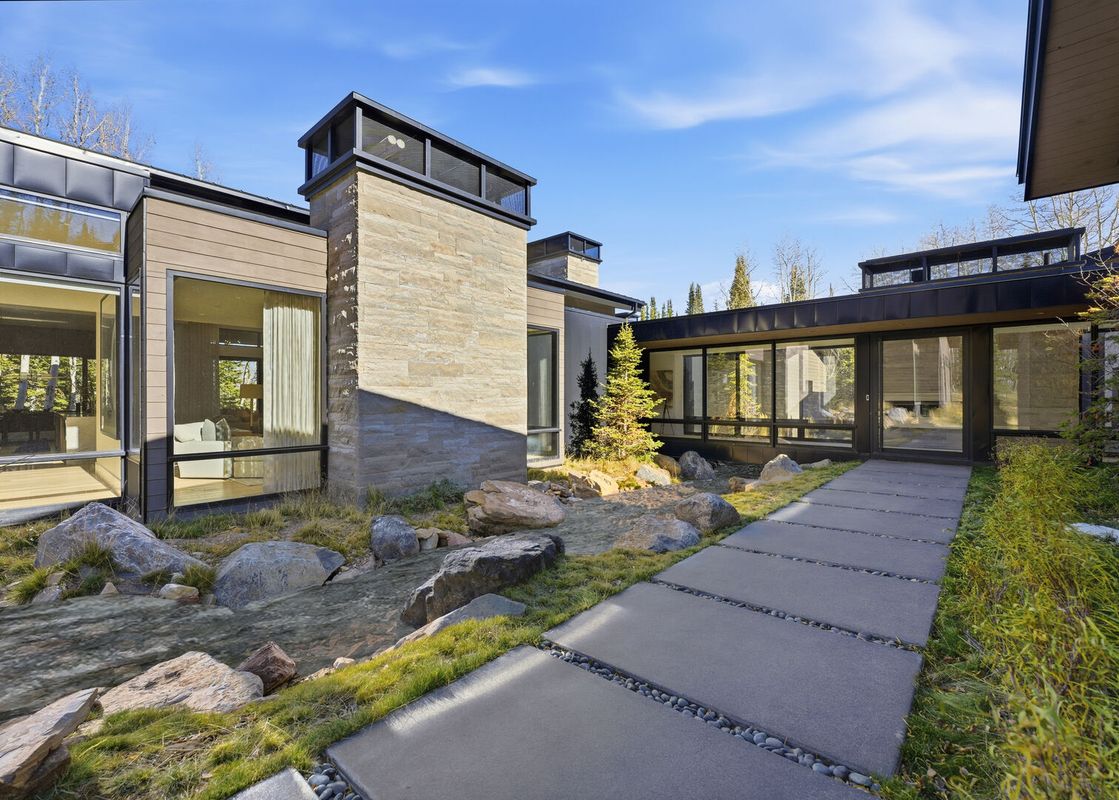 The exterior of Kestrel at the Colony features modern architecture, large windows, a rock-lined stream, and a paved walkway leading to the entrance.