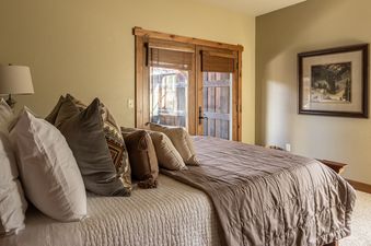 A cozy bedroom with a large bed, multiple pillows, a bedside lamp, and wooden french doors leading to an outdoor area.