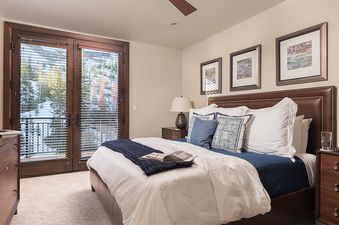 A cozy bedroom at Over the Edge features a king-sized bed with white and blue bedding, dark wooden furniture, and glass doors leading to a balcony with a snowy mountain view.