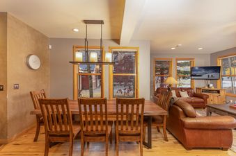 A spacious dining room with a large wooden table and chairs is connected to a comfortable living area with leather sofas and a TV.