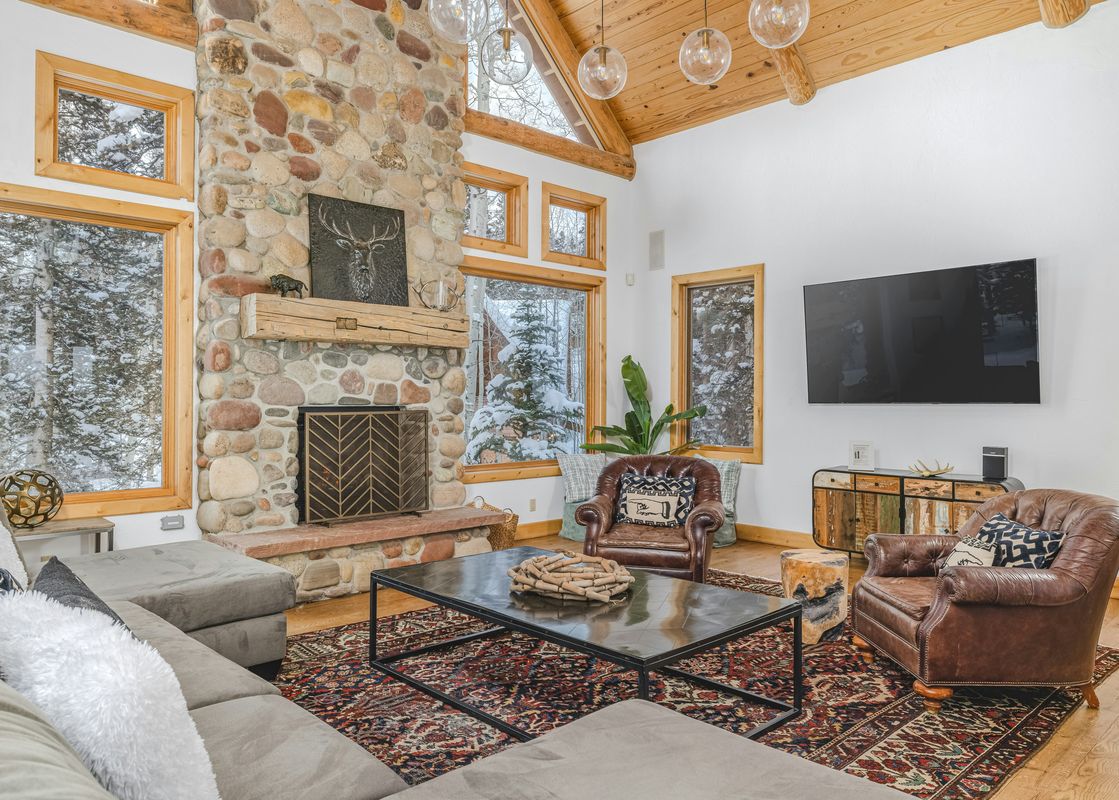 A cozy living room in Palmyra 94 Mountain Tranquility features a stone fireplace, comfortable seating, and large windows looking out onto a snowy landscape.