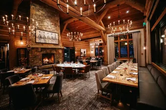 A rustic-chic dining area at The Lodge Cabins Four Bedroom Meadow Cabin features multiple dining tables set with dinnerware, large chandeliers, a stone fireplace, and log cabin walls, exuding a warm and inviting atmosphere.