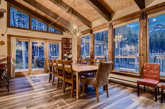The dining area at Breck Lodge features a large wooden table and chairs, surrounded by expansive windows that offer stunning views of the snow-covered mountains and dense pine forest.