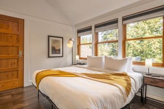 A king bedroom at Cedar Crest Eagle Cottage 8 with a white bed, yellow throw, and large windows looking out to green trees.
