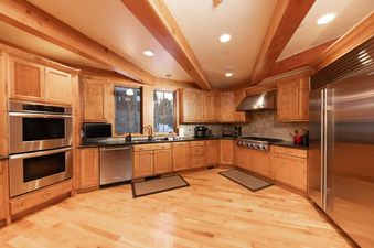 A spacious and well-lit kitchen featuring light wood cabinetry, black countertops, and stainless steel appliances, with large windows overlooking a winter landscape.