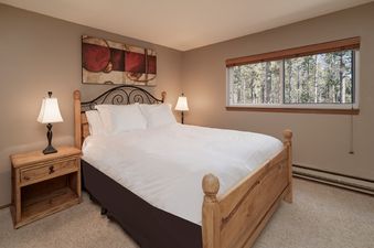 The bedroom features a comfortable queen-size bed, two bedside tables with lamps, and a large window overlooking a forest.
