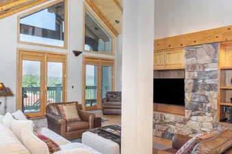 A spacious living room with a large stone fireplace, a television, and a white sofa with pillows, a view of trees outside through a large window.