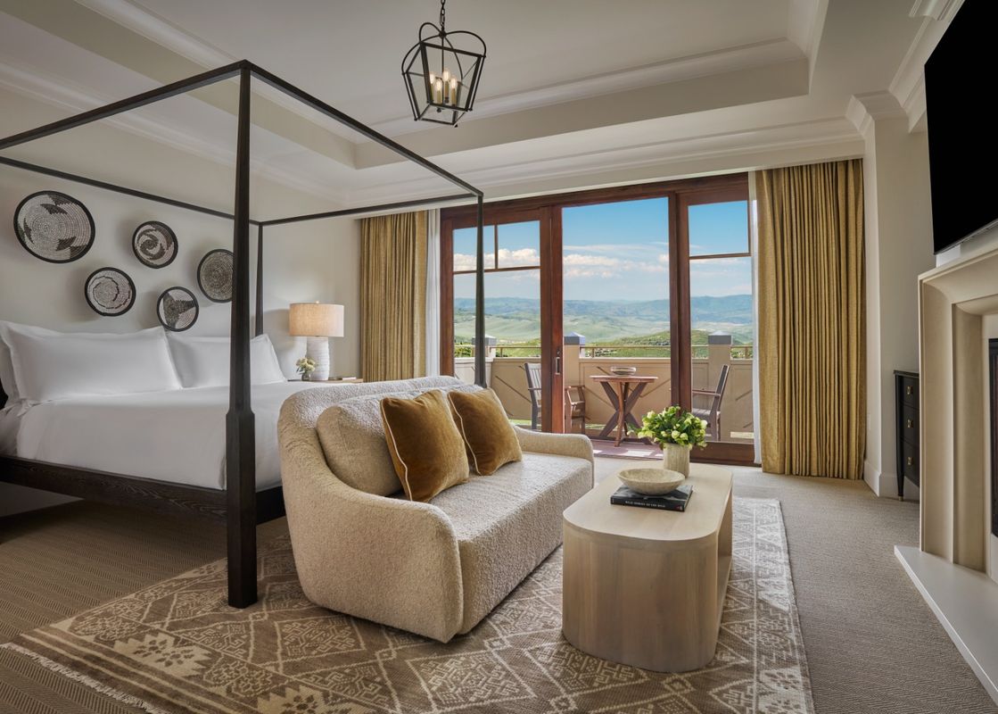 The primary bedroom features a large, comfortable bed with an elegant dark wooden frame, and a cozy fireplace, creating an inviting and warm ambiance. A spacious balcony is visible through the doors, offering picturesque views of the outdoors.