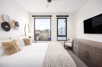 A modern master bedroom with a king-sized bed, decorative wall art, and large windows with a view of the surrounding buildings in winter.