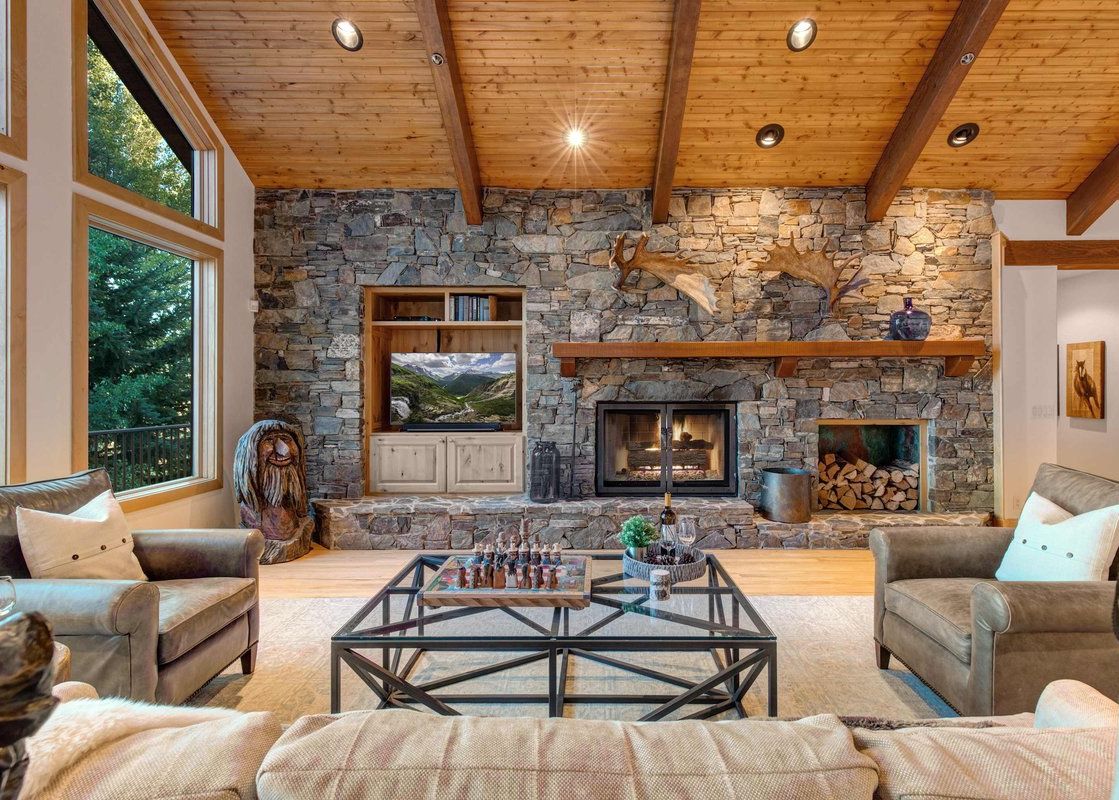 A cozy rustic living room featuring a massive stone fireplace, leather armchairs, and vaulted wood-beamed ceilings.