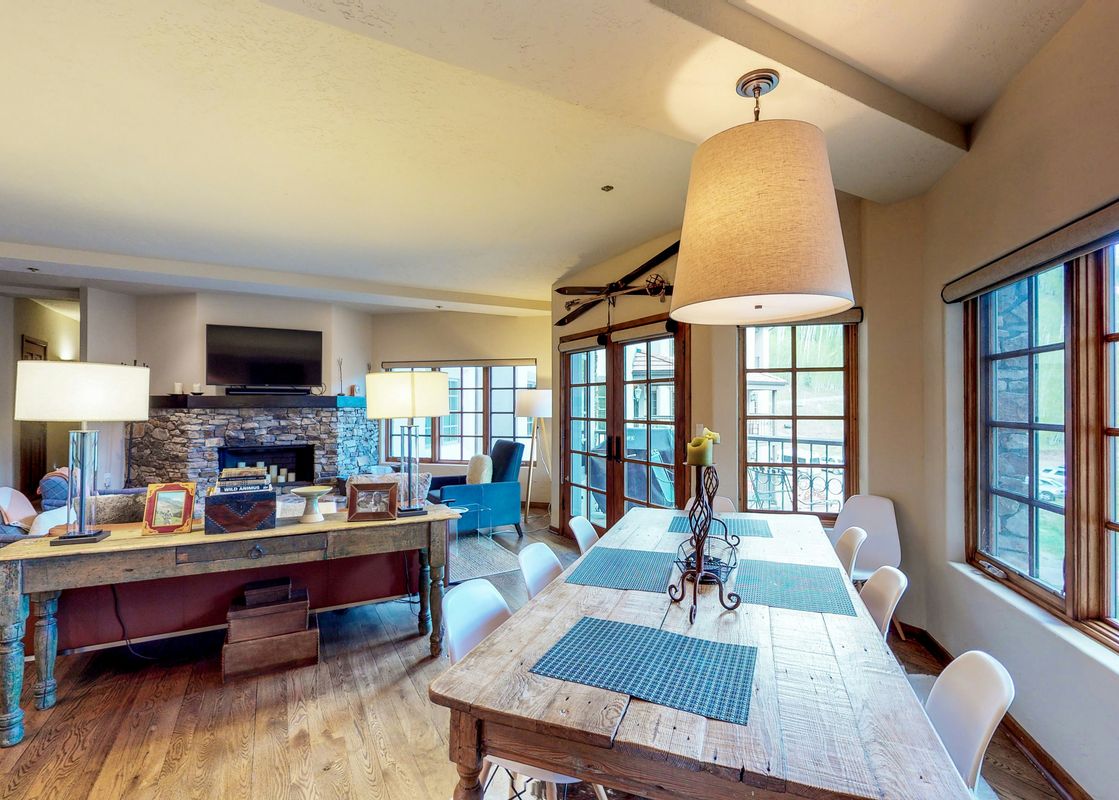 The dining area of Westermere 310 features a rustic wooden table with blue placemats, a chandelier, and a view of the mountains through the windows.