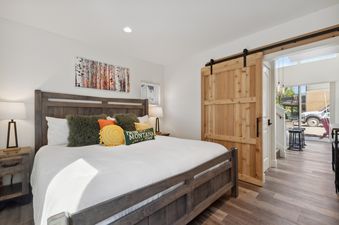 A cozy bedroom at Quarry A21 with a dark wooden bed frame, white comforter, colorful pillows, and a rustic wooden barn door leading to another area.