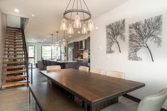 The dining area and kitchen of Riverside Reserve, featuring a long wooden dining table, modern chandelier, and dark-toned kitchen cabinetry with stainless steel appliances, all within an open-concept living space.