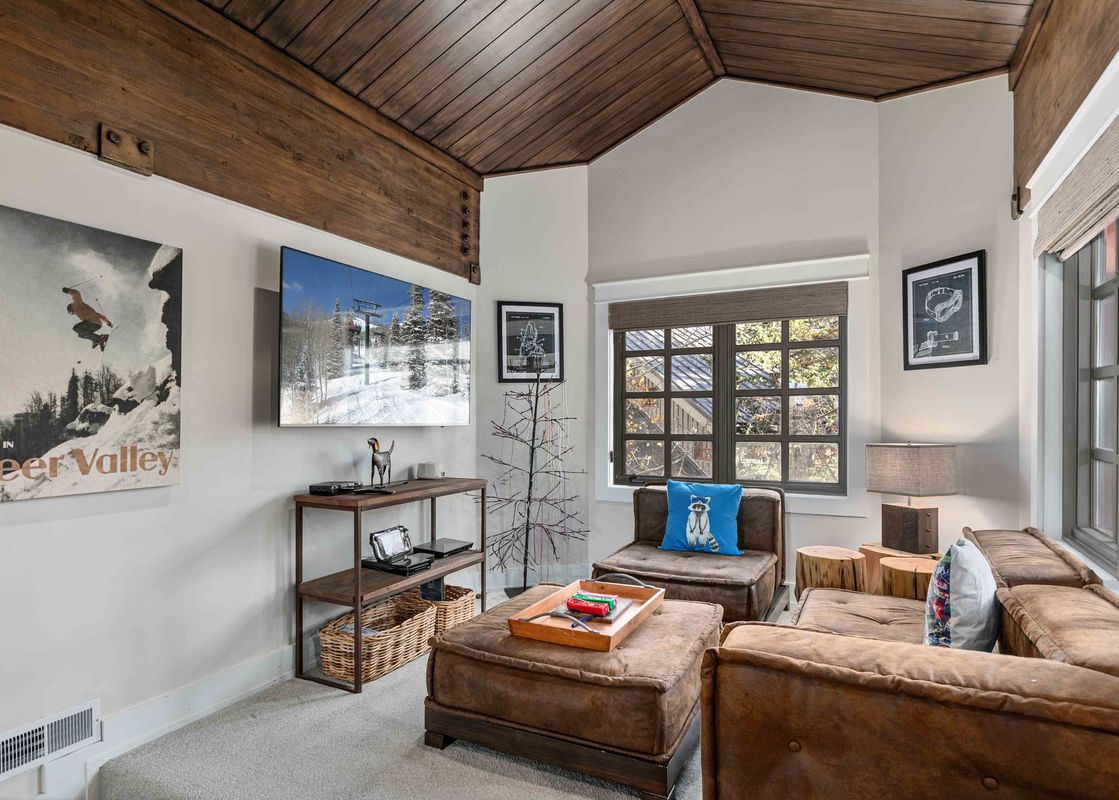 A cozy living room with a large TV, comfortable brown leather seating, a rustic wooden beamed ceiling, and a window with an outdoor view.