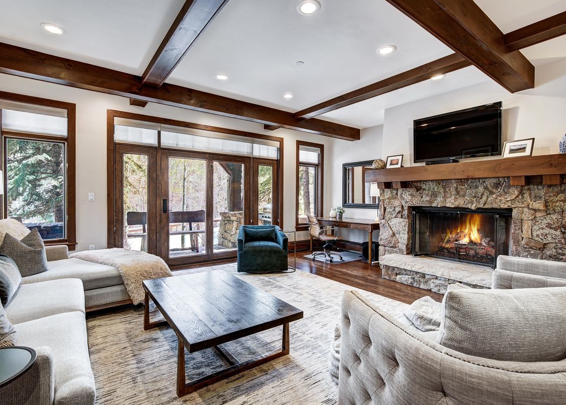 A cozy living room with a large stone fireplace, plush sofas, and a view of snow-covered evergreens through large windows and a glass door.