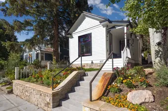 The exterior of Magic on Main features white siding, a black-framed window, and a terraced garden with colorful flowers, all accessible by concrete stairs with black railings.