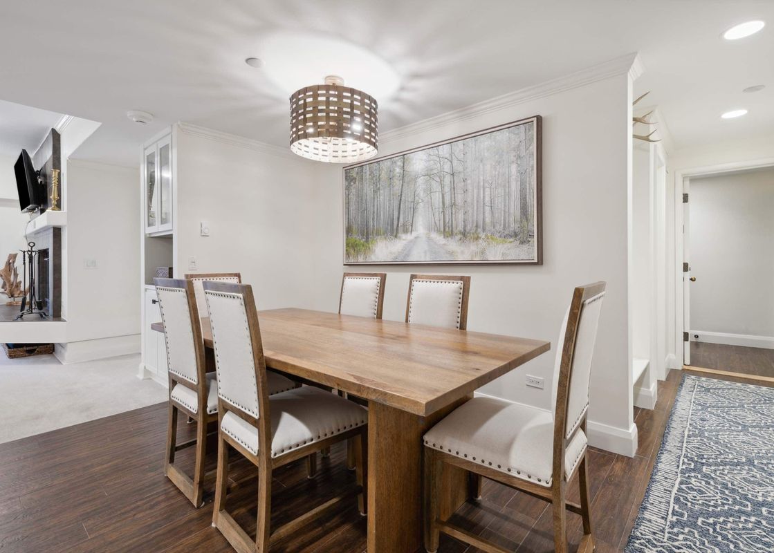 A dining area in The Charter C130 featuring a large wooden table, six upholstered chairs, and a unique drum-shaped chandelier over a forest landscape painting.