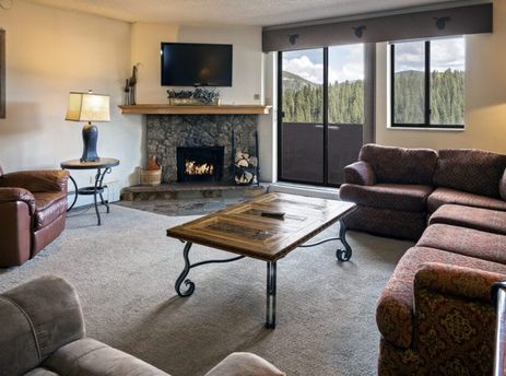 A comfortable living room at One Bedroom Suite Beaver Run Resort features a stone fireplace, a large sectional sofa, a brown armchair, and a coffee table, with glass doors leading to a balcony with mountain views.