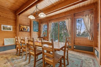 A rustic dining area with a long wooden table, eight wooden chairs, and large windows looking out onto a forested landscape.