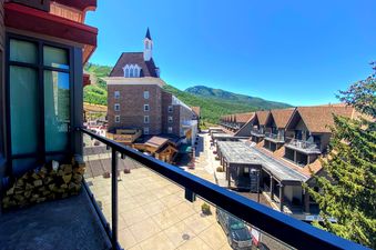 A vibrant summer day view from a balcony at The Lowell 401, overlooking a village with shops and a mountain landscape under a clear blue sky.