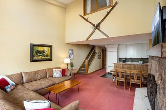 The living room at Montaneros 417 featuring a comfortable L-shaped leather sectional sofa, a wooden coffee table, a red patterned carpet, and a kitchen with white cabinets and a breakfast bar.