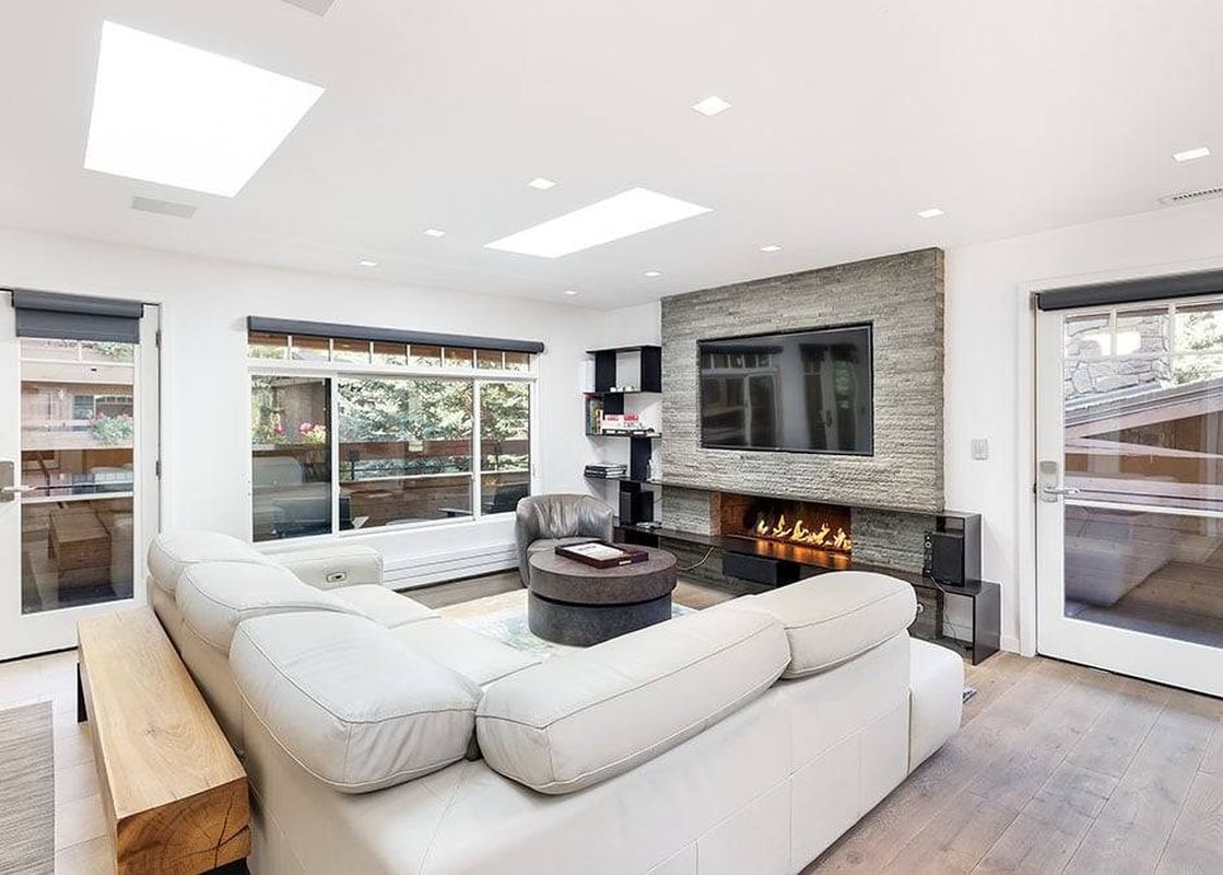 The modern living room of Fasching Haus Unit 390 features a large white sectional sofa, a stone fireplace with a mounted TV, and expansive windows offering outdoor views.