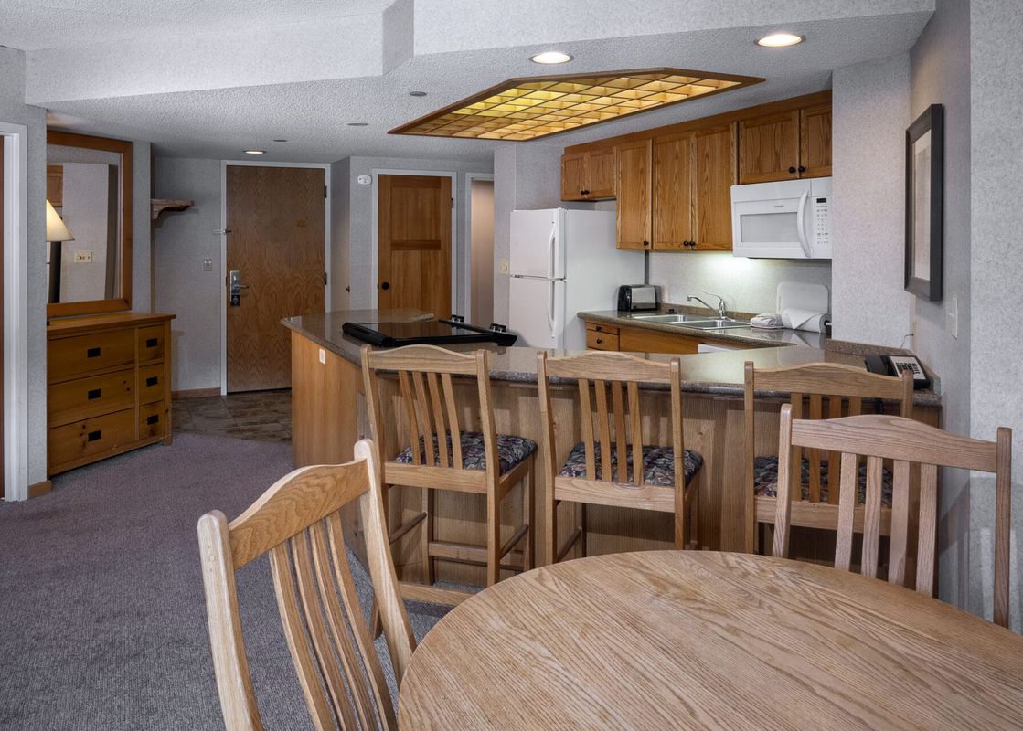 A fully equipped kitchen with wooden cabinets, granite countertops, and a breakfast bar with stools, adjacent to a round dining table in Building 4 Two Bedroom Breckenridge Suite.