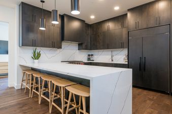 A modern kitchen with white countertops and dark wood cabinets, four wooden bar stools are lined up at the island.