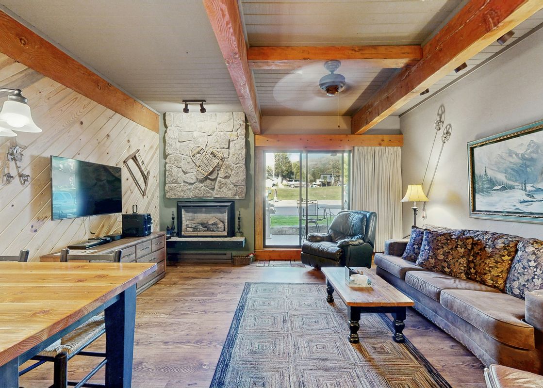 The cozy living area of Storm Meadows Club A216 showcases a stone fireplace, a flat-screen TV, and comfortable seating, with wooden accents and an outdoor view through a sliding glass door.