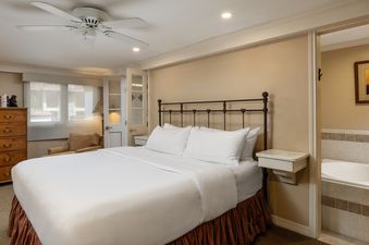 A cozy bedroom in Montaneros 406 features a king-sized bed with white bedding, a wooden dresser, an armchair by the window, and a visible bathtub through an open doorway.