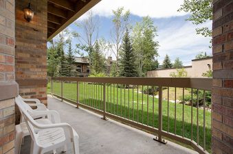A private patio with two white chairs overlooks a green lawn and trees, enclosed by a metal railing, under a bright sky.
