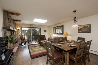 A cozy living area features a stone fireplace, a dining table set for six, and a comfortable seating arrangement with a view of the outdoors.
