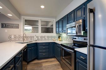 A modern kitchen at Chateau Chamonix CX113 features dark blue cabinets, white countertops, stainless steel appliances, a patterned backsplash, and wood-patterned flooring.